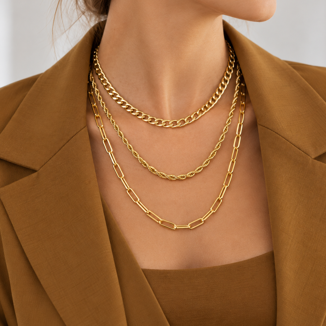 A close-up of a model wearing three layered gold-tone chains: a delicate curb chain, a twisted rope chain, and an elongated paperclip chain, styled with a brown blazer.